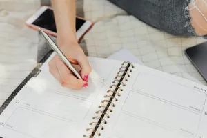 A hand with pink nail polish holds a silver pen, writing in a spiral-bound weekly planner open to a July page, with a pink phone and a dark tablet resting nearby on a light checkered blanket.