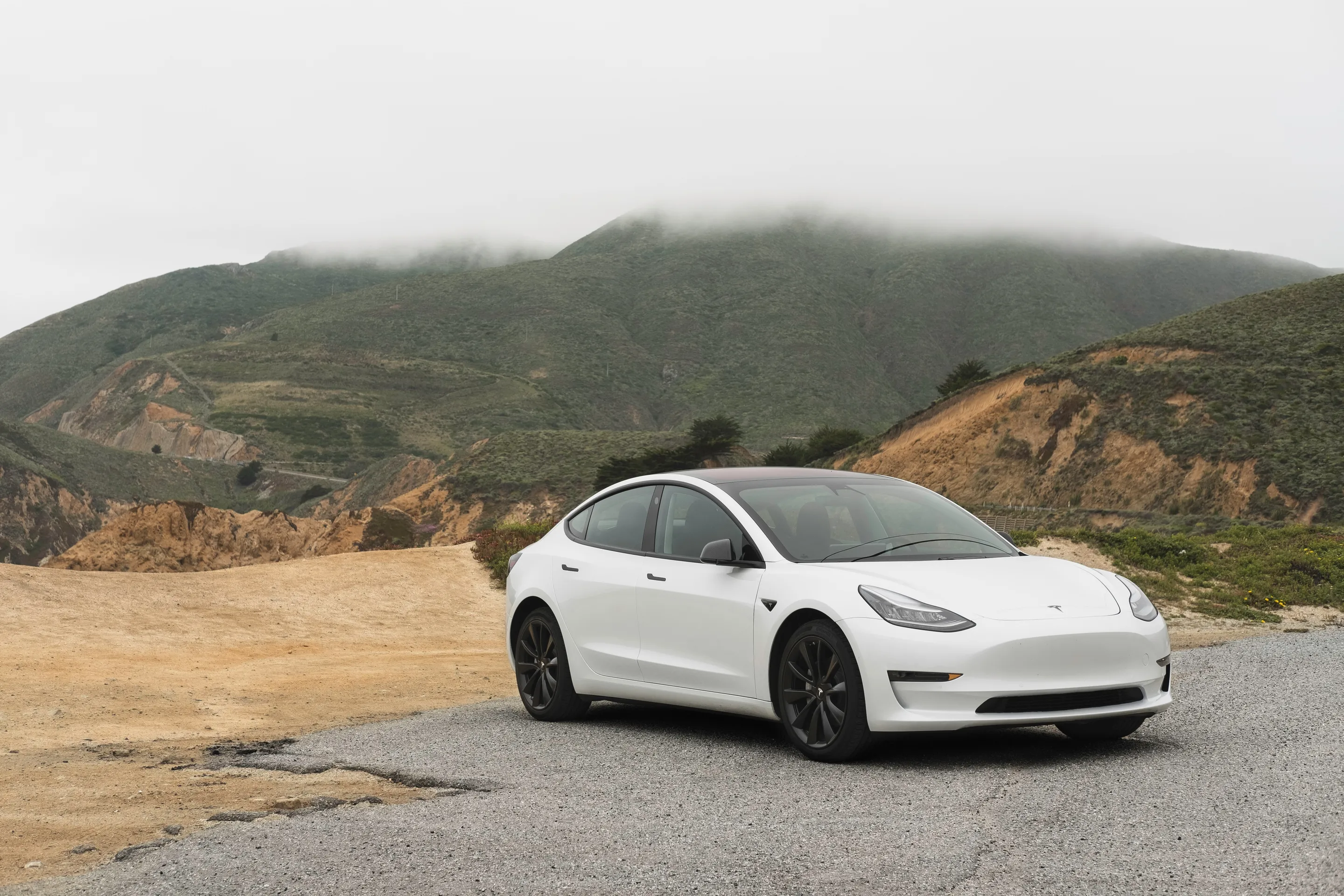 A white Tesla Model 3 sedan parked on a gravel turnout with fog-covered green mountains in the background.