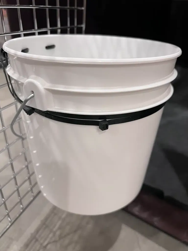 Side view of the water bucket zip-tied to the crate's wire wall, showing the mounting angle from outside the crate.