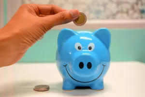A hand drops a gold coin into a smiling blue piggy bank, with a silver coin already resting on the table beside it.