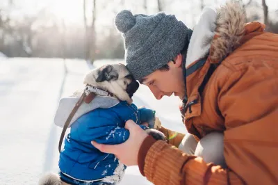 A man in an orange winter coat and gray beanie touches foreheads with a pug wearing a blue jacket, both bundled up outdoors in the snow.