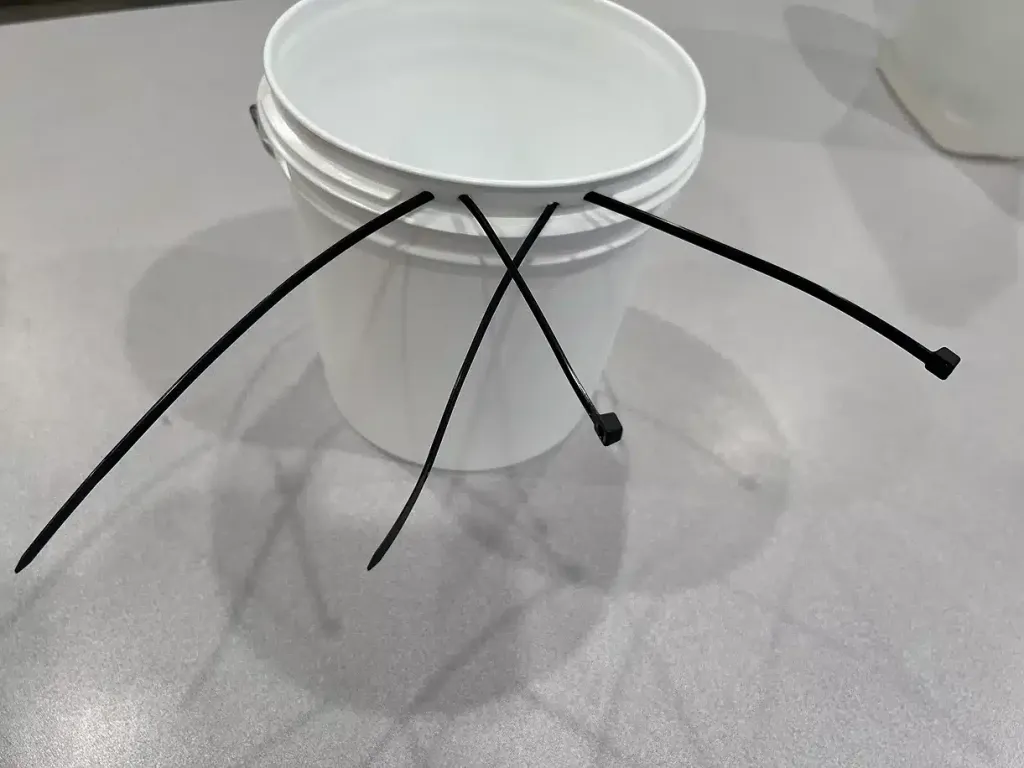 The same white 5-gallon bucket with four black zip ties threaded through the rim holes, staged and ready to be fastened inside a travel crate.