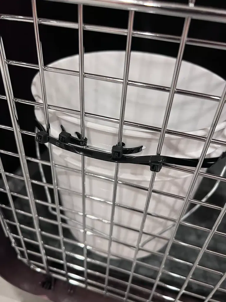 Overhead view looking down through a wire crate door at the water bucket zip-tied to the crate's interior grid.