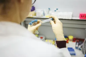 A gloved lab worker in a white coat holds several blue-capped sample tubes over a lab counter, viewed from behind over their shoulder.