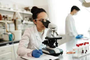 A woman in a white lab coat, safety goggles, and blue gloves looks through a microscope and takes notes, with labeled blood sample tubes on the counter and another masked lab worker in the background.