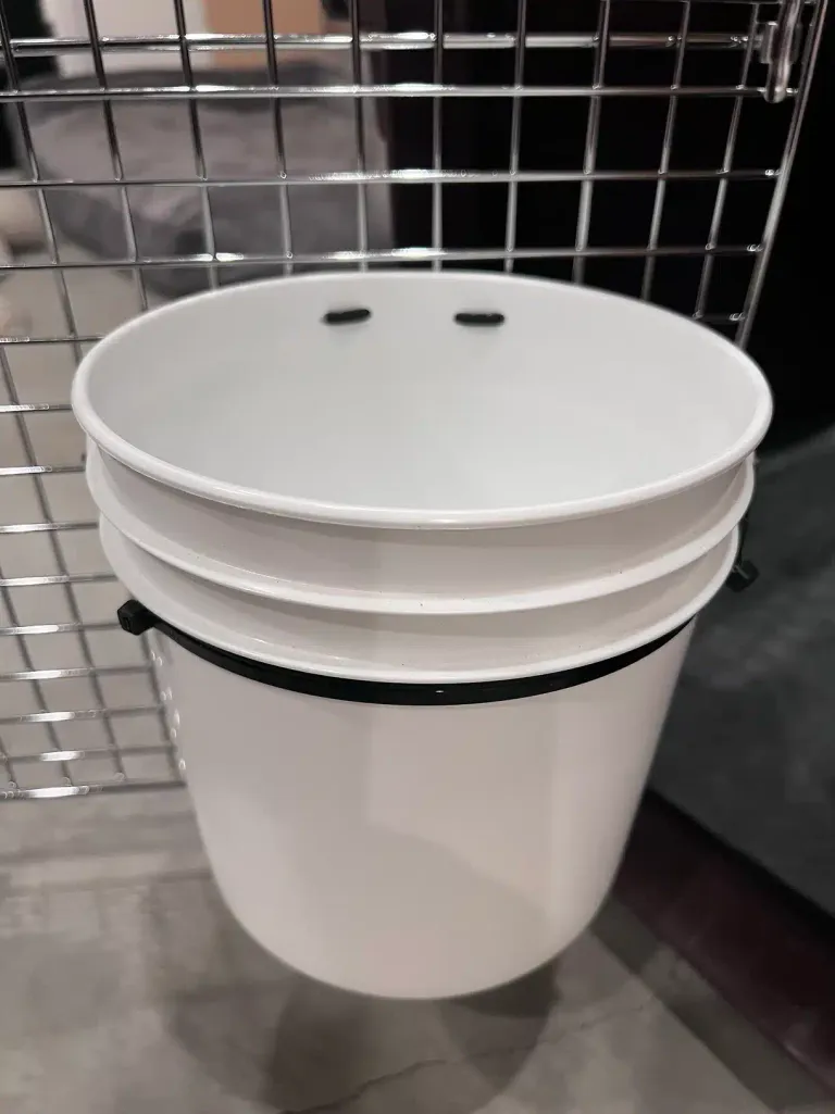 Front view of the water bucket mounted inside the wire crate, held to the crate wall by a black zip tie wrapped around the bucket's rim.