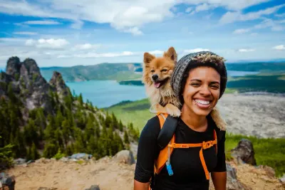A smiling woman wearing a hiking backpack and knit beanie has a small tan long-haired dog perched on her shoulder, with a mountain lake and forest in the background.